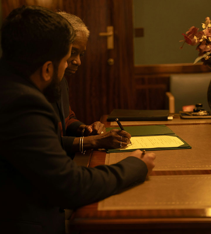 Two people sit at a wooden desk, reviewing and signing a document together. One person holds a pen over the paper while the other observes. The setting appears to be a formal office or meeting room.