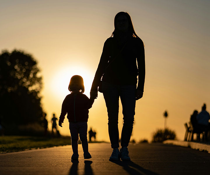 Silhouette of an adult and a child holding hands, walking on a path at sunset. The sun is low in the sky behind them, casting a warm golden glow. Other people are visible in the distance.