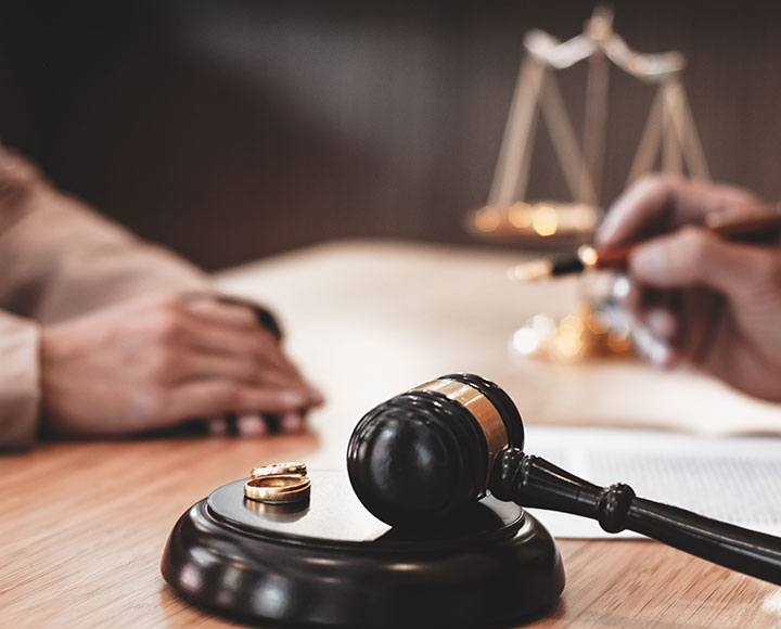 A judge’s gavel and two wedding rings rest on a desk, while two people sit across from each other, one writing on a document; a blurred scale of justice is in the background.