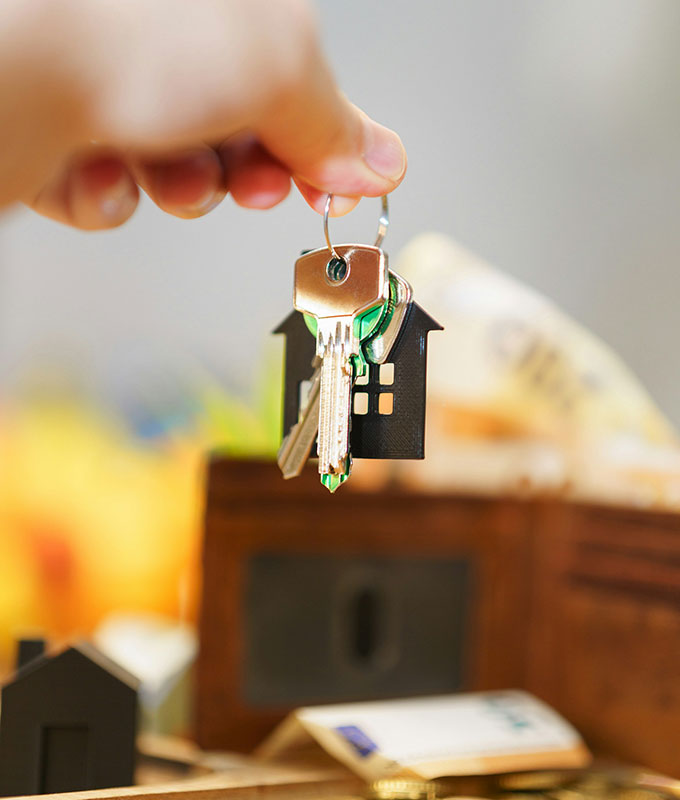 A hand holding a keyring with several keys and a black house-shaped keychain, with a blurred background featuring a wooden box and other indistinct items.