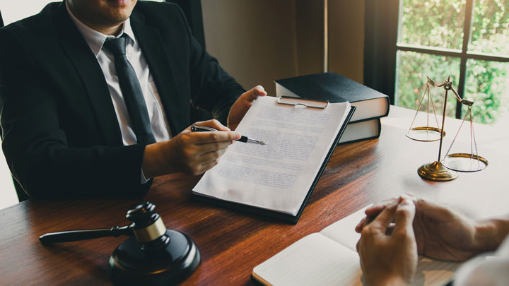 A lawyer in a suit points to a document on a clipboard while discussing with a client. Legal books, a gavel, and scales of justice are on the wooden desk near a window.