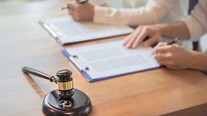 A gavel and two wedding rings rest on a wooden table in the foreground, while two people in the background fill out documents on clipboards, suggesting a legal or divorce proceeding.