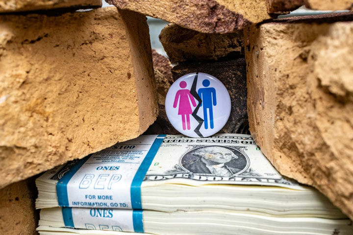 A round pin with stick figures of a man and woman stands upright among bricks above stacks of one-dollar bills.