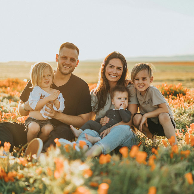 A family of five sits together in a field of orange flowers, smiling at the camera. Two adults and three young children are surrounded by vibrant blooms under bright sunlight.