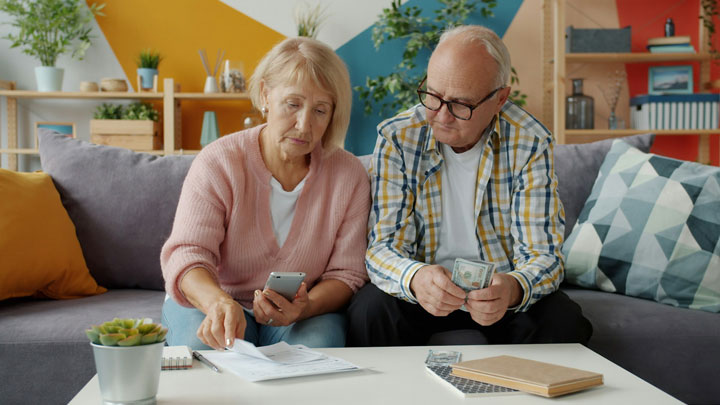 An elderly couple sits on a couch, reviewing documents and using a smartphone. The woman points at papers on the table, while the man holds a stack of cash. The room has colorful decor and plants in the background.