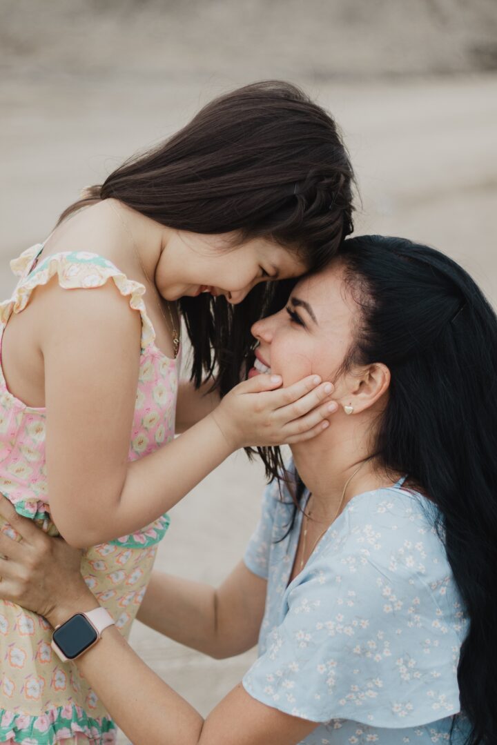 A young girl and a woman smile warmly at each other, touching foreheads. The woman kneels, holding the girl, and they both look happy and affectionate outdoors.