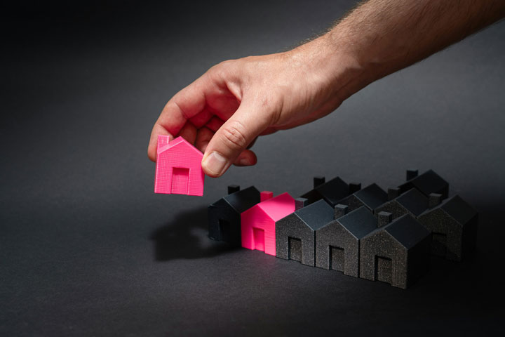 A hand picks up a small pink house model from a group of black house models on a dark surface, highlighting the pink houses among the others.