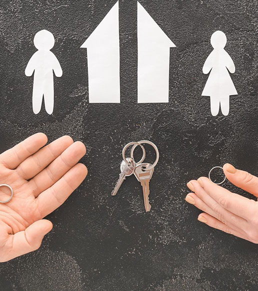 Two hands face each other, each holding a wedding ring. Paper cutouts of a man, a woman, and two houses are above, with a set of house keys between the hands, symbolizing separation or divorce.