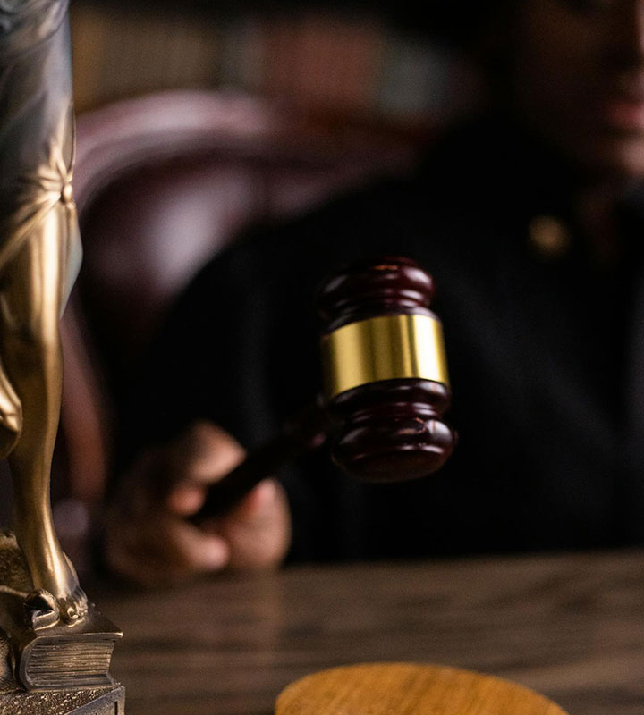 A judge's hand holding a wooden gavel poised above a desk, with a blurred statue and legal books in the background.