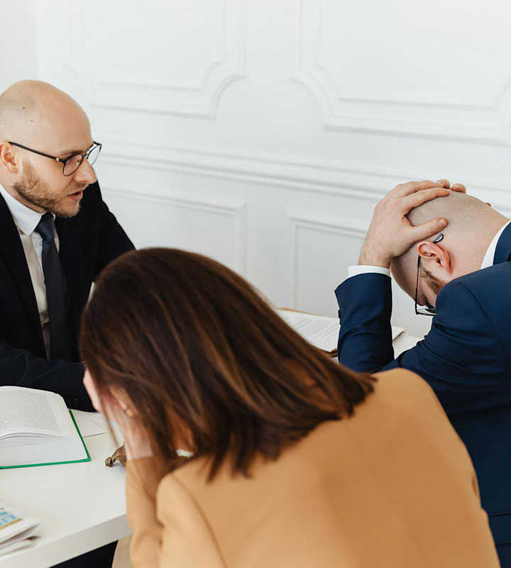 Three people sit at a table in an office. Two men in suits, one with his head in his hands appearing stressed, while the other talks. A woman in a tan blazer sits with her head down, also looking upset.