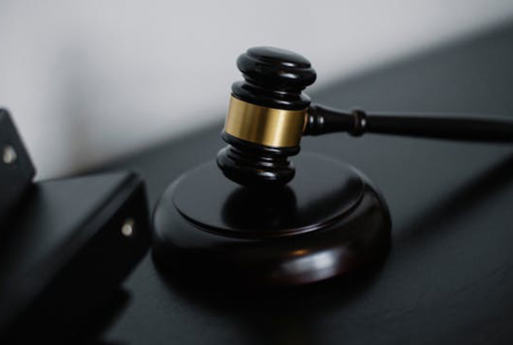 A close-up of a wooden judge’s gavel with a gold band resting on a sound block, symbolizing law and justice, on a dark surface with part of a folder or book visible next to it.