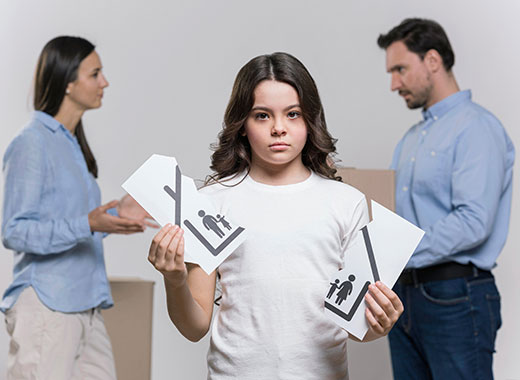 A sad girl stands holding two torn pieces of paper with family symbols, while a man and woman argue in the background, suggesting a family conflict or divorce.