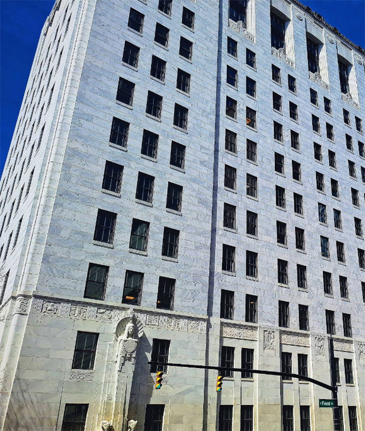 A tall, white stone building with many windows stands against a clear blue sky. Decorative carvings adorn the lower facade above the entrance. A traffic light hangs above the street in front of the building.