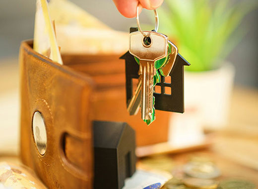A hand holds house-shaped keychains with keys in front of an open brown wallet filled with cash, coins, and a small house model, symbolizing real estate or home buying.