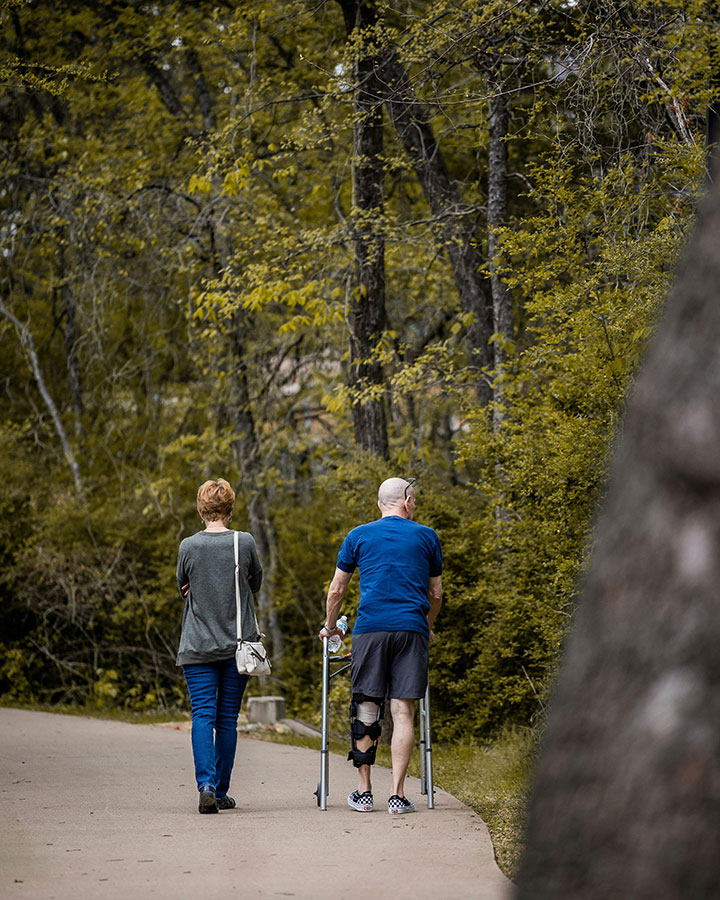 A woman and a man with a knee brace and crutches walk along a paved path surrounded by trees and greenery. The pair are seen from behind, enjoying a peaceful outdoor setting.