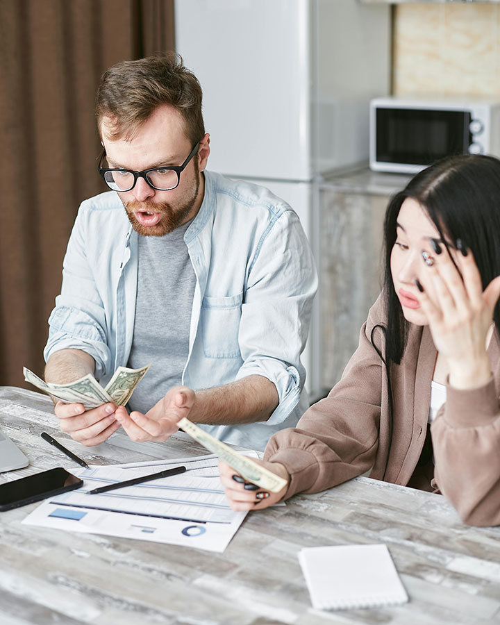 A man and woman sit at a table with papers, a laptop, and cash, both looking stressed and worried as they count money and review documents.
