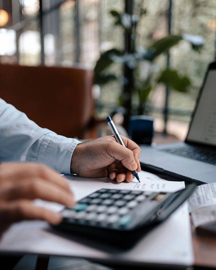 A person uses a calculator and writes on a piece of paper at a desk with a laptop, indoors with natural light and greenery visible through large windows in the background.