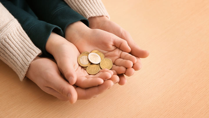 Close-up of adult hands gently holding a child's cupped hands, which are holding several coins, on a light wooden surface.