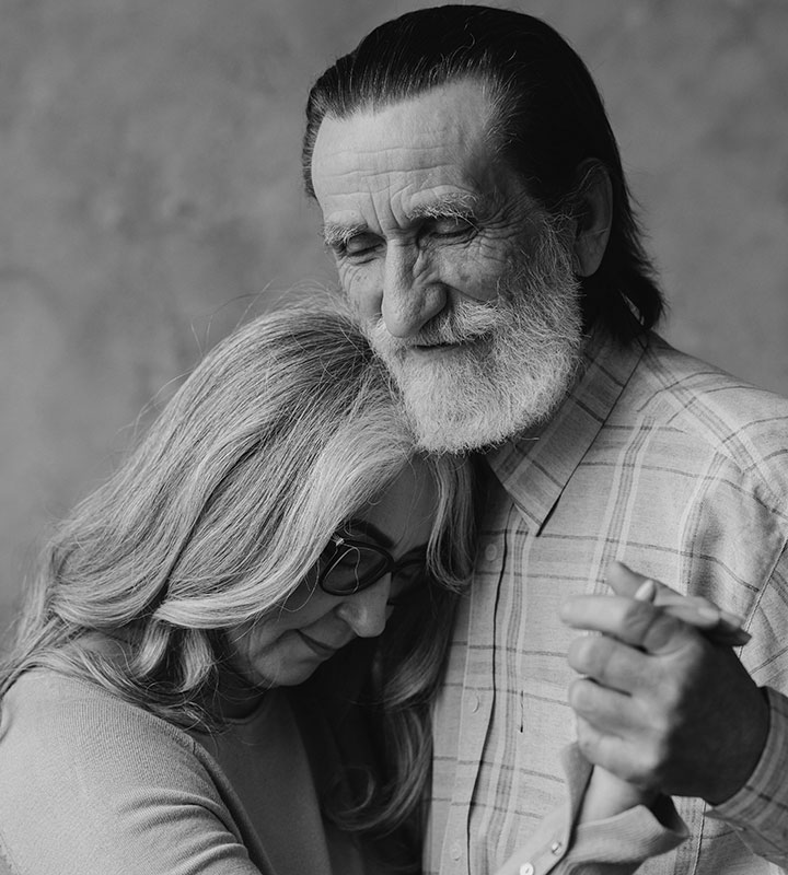 An elderly couple embraces while dancing. The man, with gray hair and a beard, smiles with his eyes closed, while the woman, with long gray hair and glasses, rests her head on his chest. The image is in black and white.