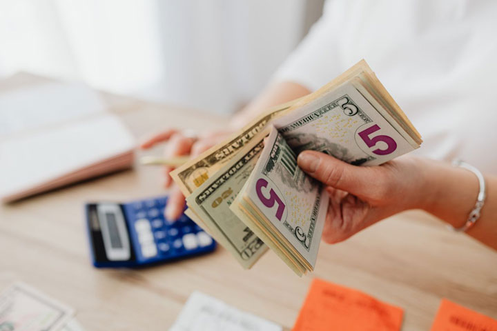 A person counts a stack of U.S. dollar bills at a desk with a blue calculator, papers, and sticky notes nearby.