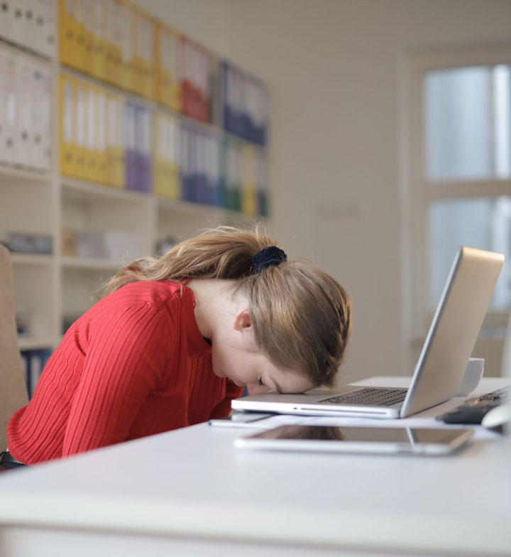 A young woman in a red sweater sits at a desk with her head resting on her open laptop, looking tired or frustrated. Shelves with binders are visible in the background.