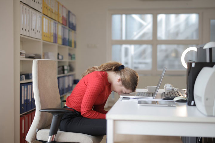 A woman in a red sweater sits at a desk with her head resting on her laptop keyboard, appearing frustrated or exhausted, in a brightly lit office with shelves of files in the background.