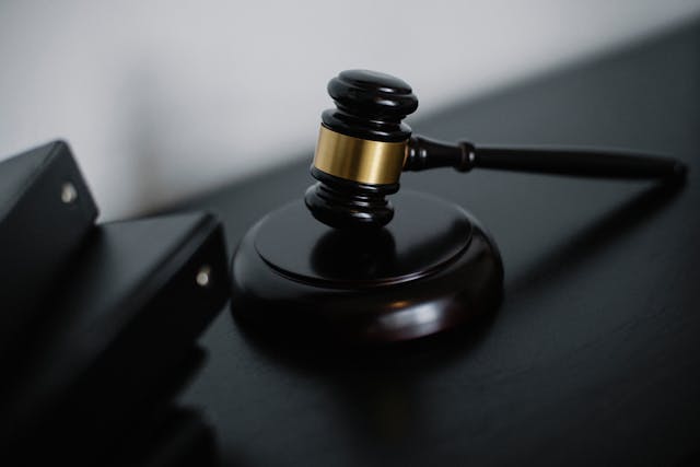 A judge’s wooden gavel with a gold band rests on a sound block on a dark table, next to a stack of black binders.