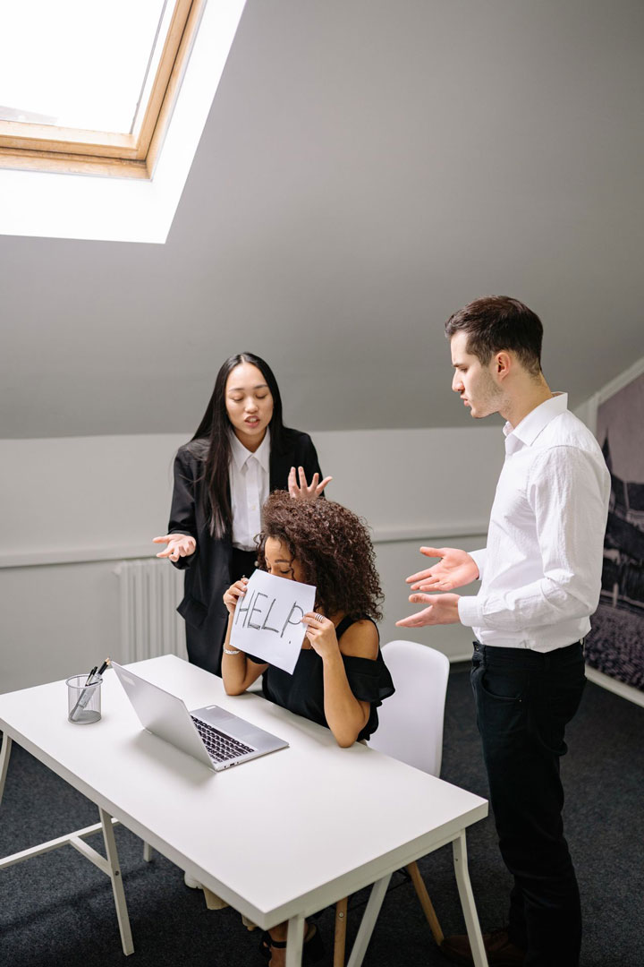 A woman sits at a desk holding a sign that says "HELP" in front of her face, while two colleagues stand beside her, gesturing as if arguing or discussing intensely. A laptop is open on the desk.