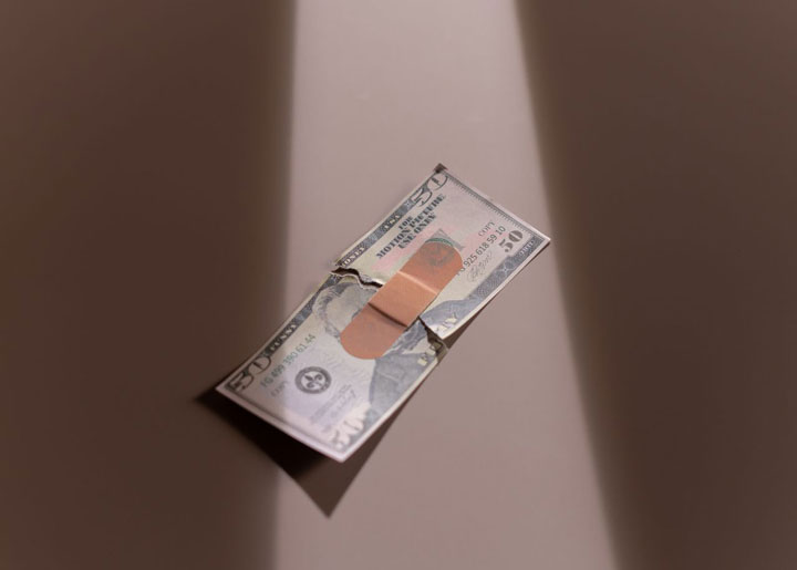 A torn $10 bill is partially held together with a bandage, lying under a beam of light on a neutral surface.