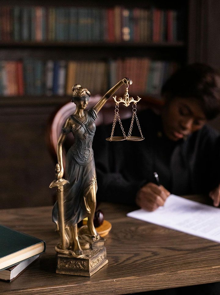 A brass statue of Lady Justice holding scales and a sword sits on a desk in front of legal books, while a person in a black robe writes on papers in the background.