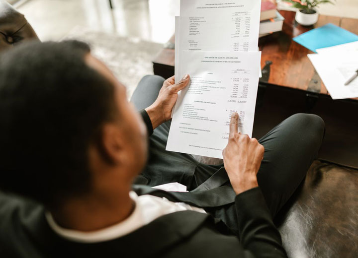 A person in a suit sitting on a couch, holding and reviewing financial documents, with one finger pointing at a section. A table with papers and a potted plant is visible in the background.