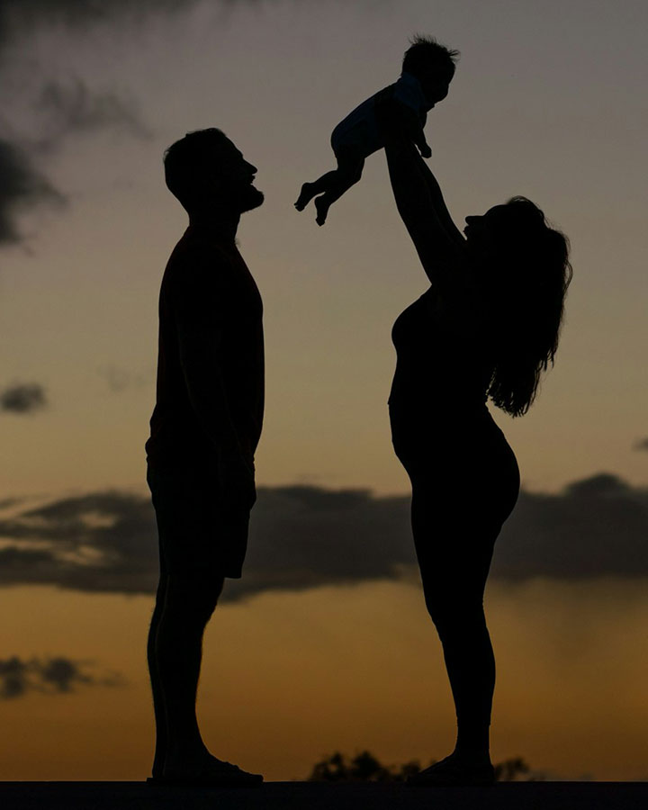 Silhouette of two adults and a baby at sunset; one adult stands facing the other, who lifts the baby joyfully overhead against a dramatic evening sky.