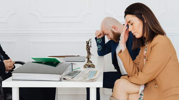 A woman and a man sit at a table looking stressed, with the woman holding her forehead and the man bowing his head. Folders, papers, and a small statue are on the table, suggesting a legal or serious discussion.