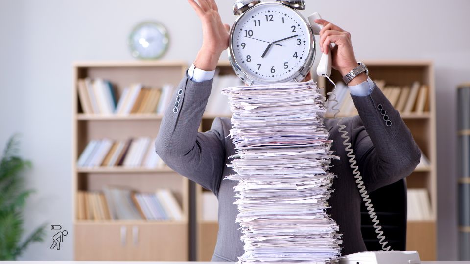 A person in a suit sits at a desk with a huge stack of papers, holding a large clock and a phone, obscuring their face. Bookshelves are in the background, symbolizing stress and time pressure at work.