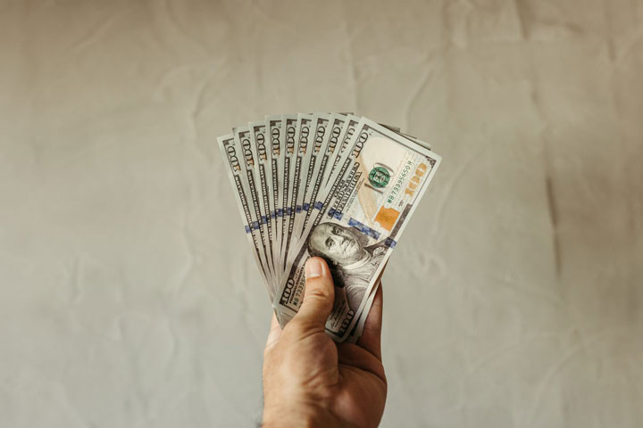 A hand holding a fanned-out stack of U.S. one hundred dollar bills against a plain, light-colored background.