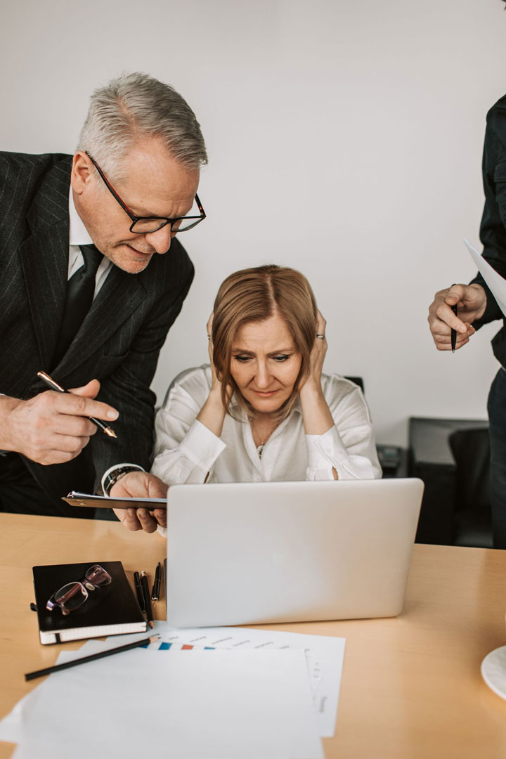 A woman sits at a desk looking stressed, holding her head, while a man in a suit stands beside her pointing at a notebook. Papers, glasses, and a laptop are on the desk. Another person stands nearby holding documents.