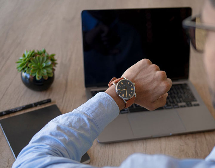 A person wearing a watch checks the time while sitting at a desk with a laptop, a notebook, a pen, and a small potted plant.