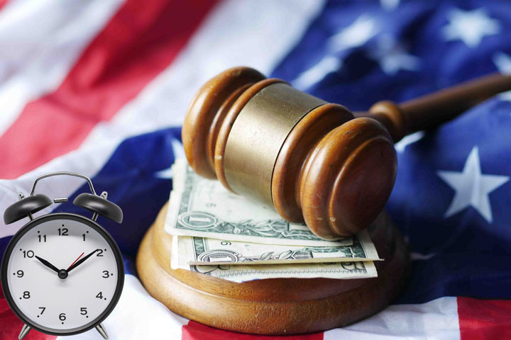 A judge’s gavel rests on dollar bills atop a folded American flag, with an alarm clock showing 10:09 positioned to the left in the foreground.