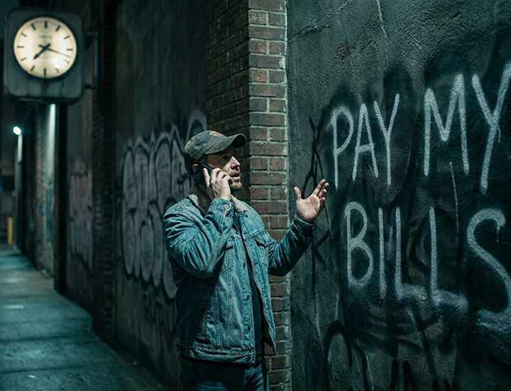A man in a denim jacket and cap talks on his phone while gesturing with his hand, standing in a dimly lit alley. Graffiti on the wall reads "PAY MY BILLS." A clock on the building shows 11:55.