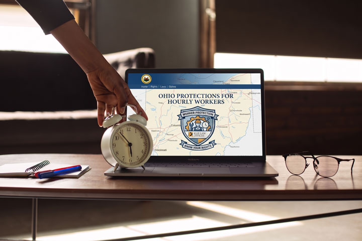 A hand adjusts an alarm clock in front of a laptop displaying a website titled “Ohio Protections for Hourly Workers.” A notebook, pen, and glasses are on the table nearby.