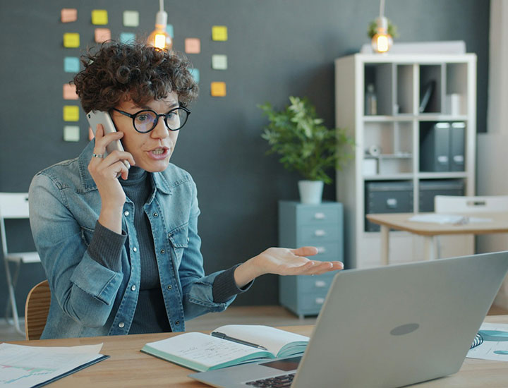 A person with curly hair and glasses, wearing a denim shirt, talks on the phone while gesturing at a laptop screen. They are sitting at a desk with notebooks and papers, with a blackboard and sticky notes in the background.