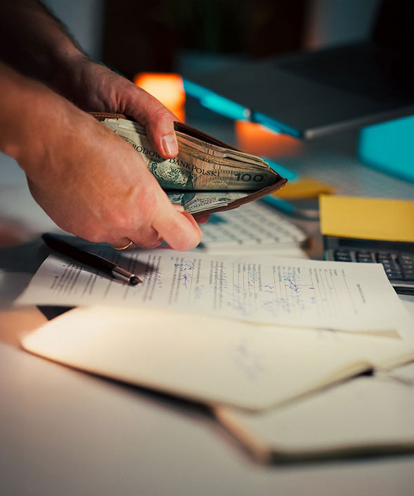 A person counts folded US dollar bills from a wallet over a desk with paperwork, a pen, a calculator, and an open notebook, suggesting financial planning or payment.