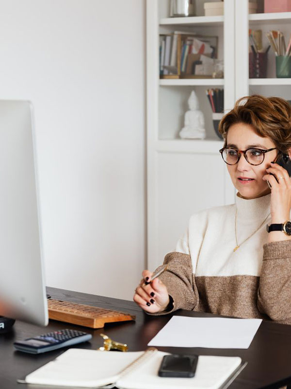 A woman wearing glasses and a sweater sits at a desk, talking on a smartphone. She holds a pen, with a computer monitor, notebook, calculator, and papers in front of her. Shelves with books are in the background.