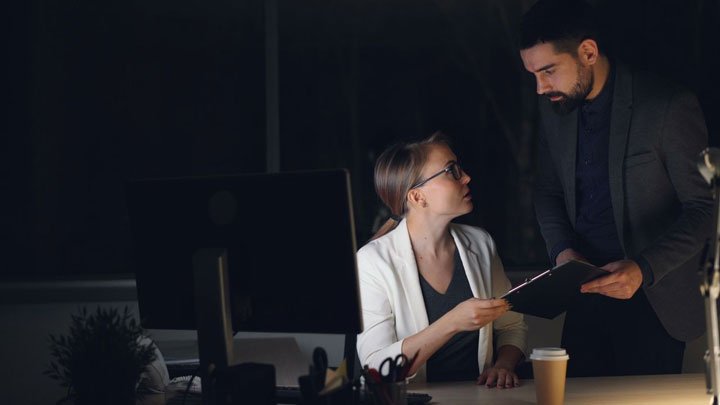 A woman sitting at a desk with a computer talks to a standing man holding a clipboard in a dimly lit office. A coffee cup and documents are on the desk.