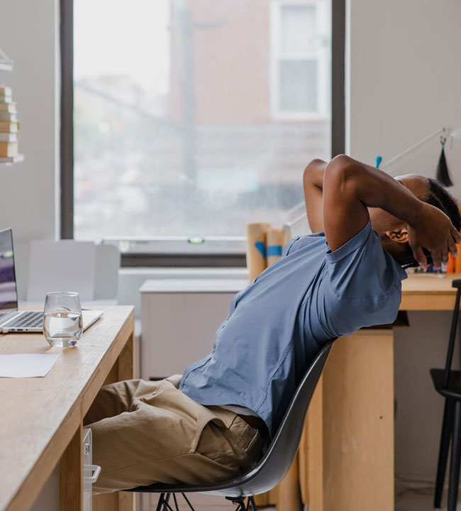 A person leans back in a chair at a desk, hands clasped behind their head, appearing relaxed or thoughtful. There is a laptop, papers, and a glass of water on the desk. A large window is in the background.
