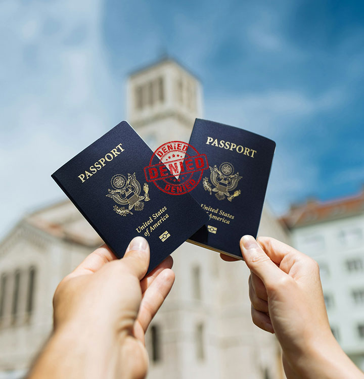 Two hands holding U.S. passports outdoors with a large red "DENIED" stamp over them; blurred buildings and a clear sky are in the background.