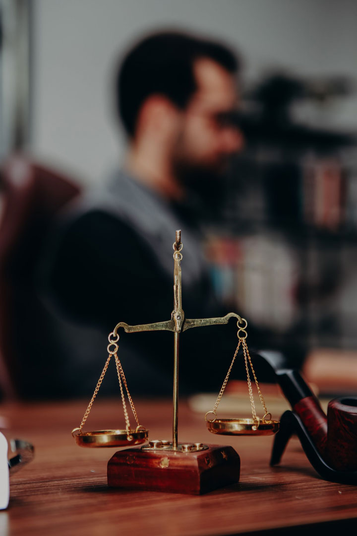 A close-up of a brass balance scale on a wooden desk, with a blurred figure of a man sitting in the background. The setting appears to be an office or study.