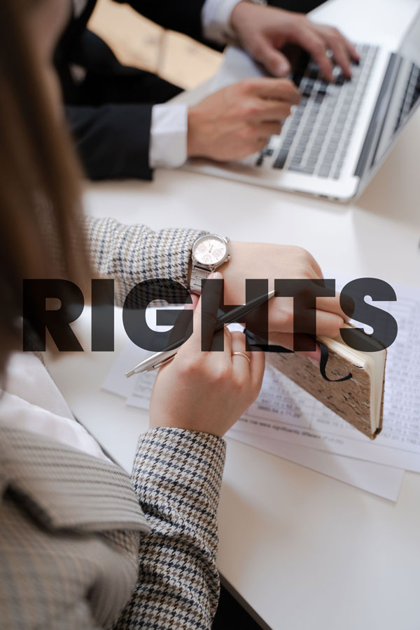 A person writing in a notebook at a desk, while another person works on a laptop in the background. The word "RIGHTS" is overlaid in bold letters across the image.