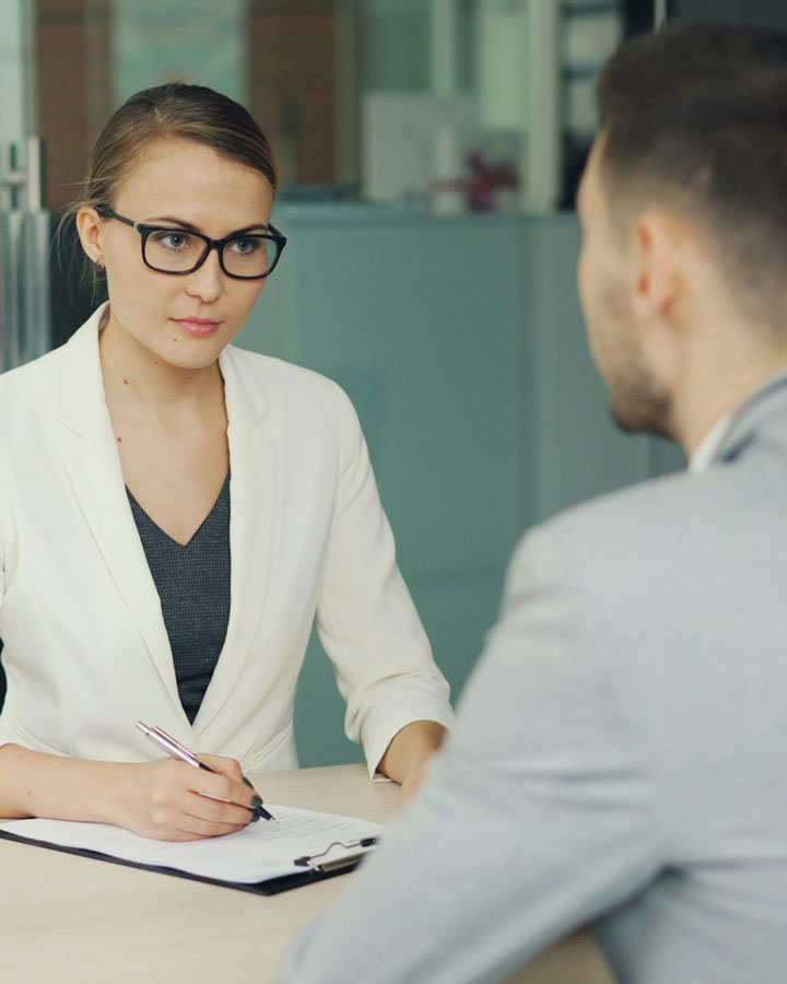A woman wearing glasses and a white blazer is sitting at a desk, holding a pen and clipboard, interviewing a man in a gray suit in an office setting.