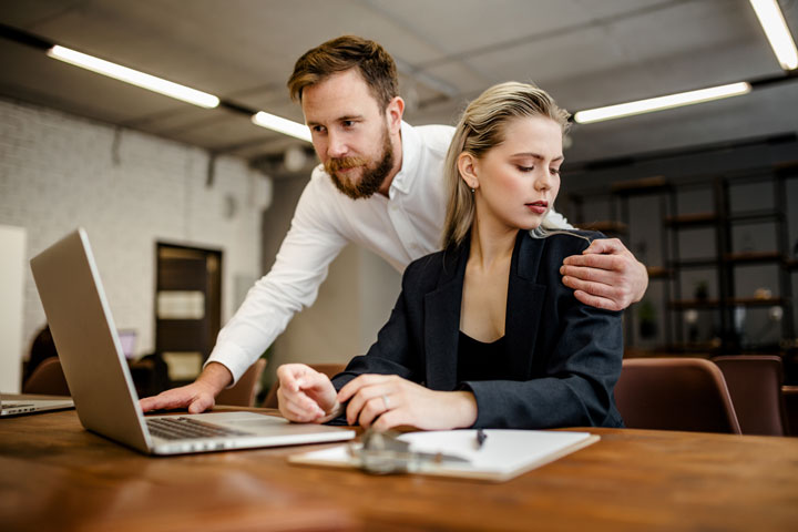 A man stands behind a seated woman in an office, placing his hand on her shoulder. The woman looks uncomfortable and turns away, while the man looks at her. A laptop and papers are on the desk.